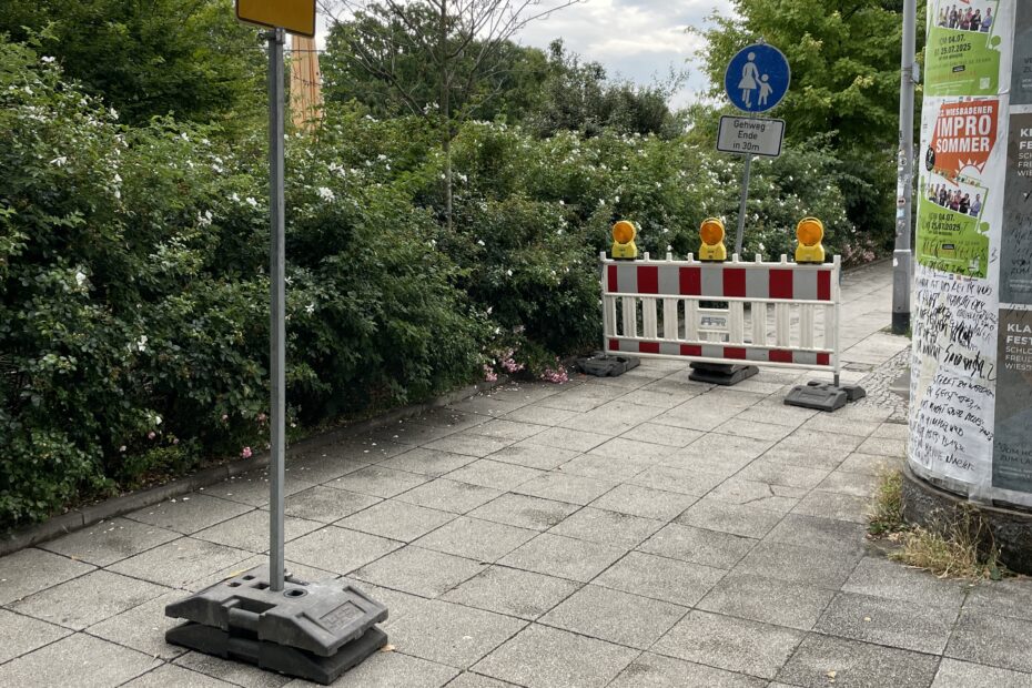 Ein Fußweg ist gesperrt. Eine rot-weiße Absperrung mit gelben Lampen blockiert den Weg. Dahinter steht ein Schild: „Gehweg endet in 30 m“. Links zeigt ein gelbes Schild mit Pfeil nach links: Fußgänger sollen hier abbiegen.