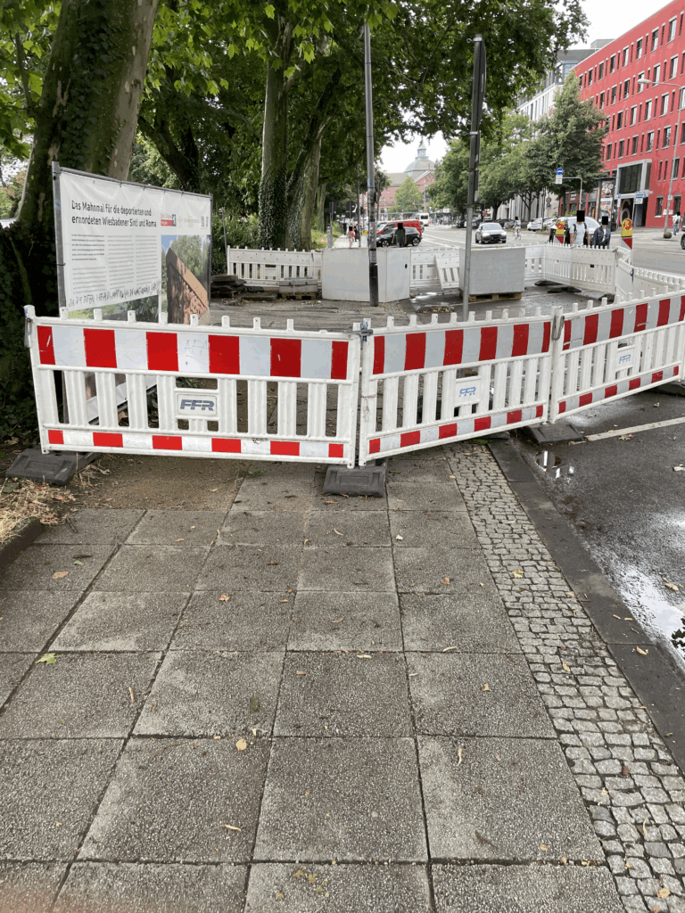 Ein Gehweg ist mit rot-weißen Baken abgesperrt. Menschen laufen auf der Straße im Hintergrund. Es gibt keinen abgesicherten Weg für Menschen zu Fuß. Dennoch gehen viele Menschen entlang der Baustelle.