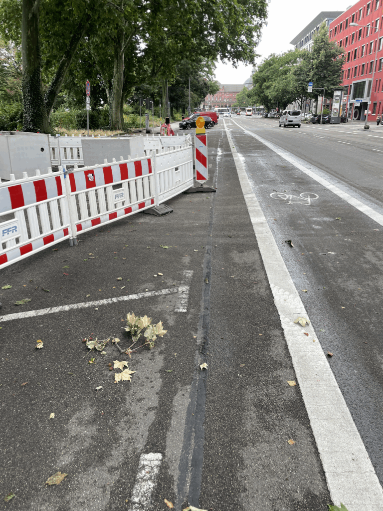 Die Baustelle ragt in einen Parkstreifen hinein. Sie ist mit rot-weißen Baken abgesperrt. Eine Warnlampe steht vorne. Daneben ist ein Fahrradstreifen.